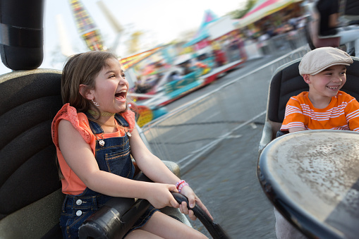 A little girl and a little boy ride a carnival ride together.  The girl holds on tight and yells, the little boy is smiling.  Both are expressing positivity and enjoyment.  Both children are Hispanic and wearing orange.  The girl has long brown hair, the boy is wearing a hat.