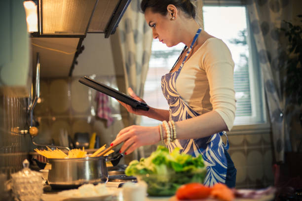 istock Young woman reading recipe on digital tablet in the kitchen and preparing spaghetti 692070626