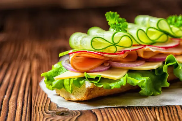 Photo of close up of sandwich with ham, cheese, bacon, radish, lettuce, cucumbers and onions on paper, wooden background