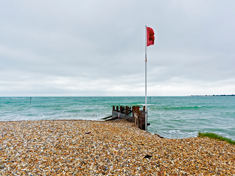On a windy West Wittering beach, under a grey sky, a red flag is flying on the shore.