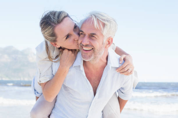 mann gibt seine lächelnden frau huckepack am strand - attractive couple stock-fotos und bilder