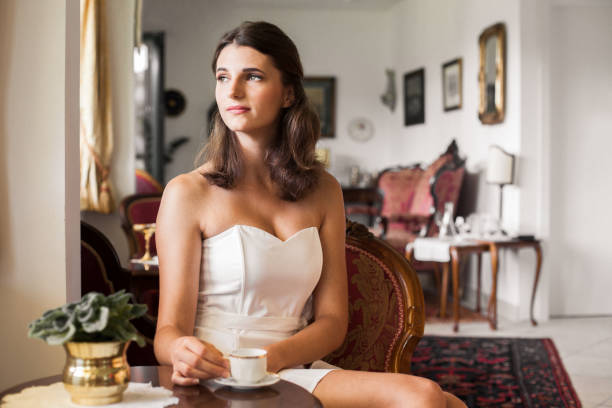 Graceful Young woman wearing a fashionable evening gown sitting in old mansion drinking a coffee, relaxing, looking out the window.