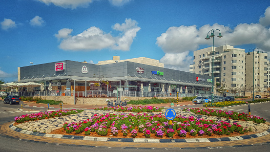 Nes Ziona, Israel-March 20, 2016:  Beautiful view of big flowerbed placed on traffic circle in front of supermarket Mega In The City and other commercial stores. Nice blue sky is over it. The complex is located at 1 Avner Ben Ner Street. Two white multi-story condominiums are seen in the background. Horizontal shot.