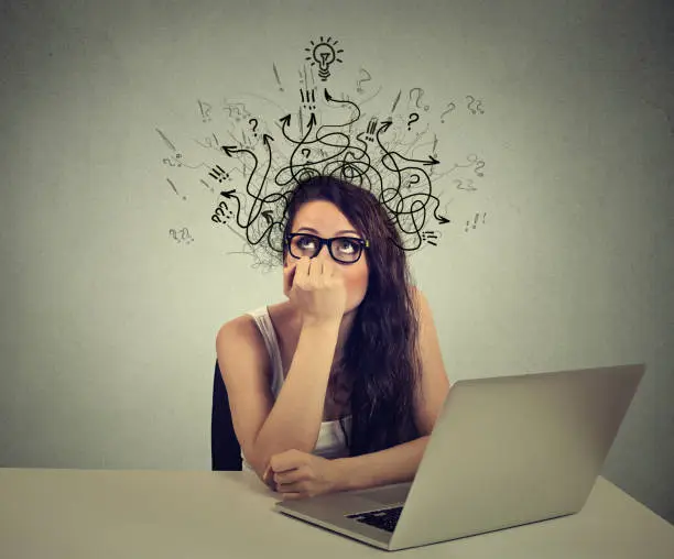 Photo of woman with thoughtful expression sitting at a desk with laptop with lines arrows and symbols coming out of her head