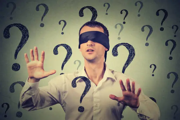 Portrait blindfolded man stretching his arms out walking through many question marks