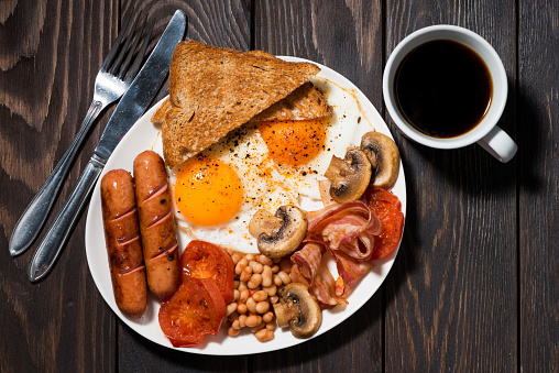 Fried eggs with bacon, sausages and vegetables for breakfast on wooden table, top view