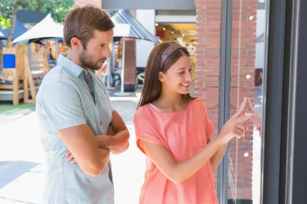 Photo of Bored man watching his happy wife looking at a window