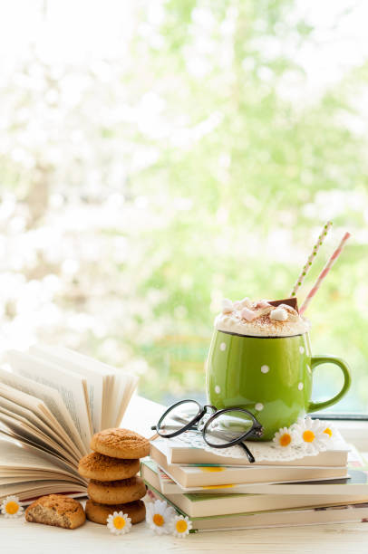 istock Coffee cup with marshmallows, open book, glasses, cookies and flowers on window with bokeh. Reading and breakfast concept 692452206