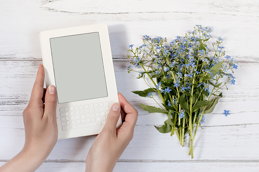 Woman hands holding white ebook, bouquet on a white rustic wooden background