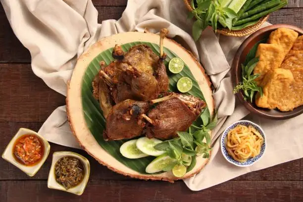 Bebek Goreng, one popular Indonesian poultry dish of deep-fried duck; served with fresh vegetables and chili paste. The duck legs are arranged on a natural wooden serving board that has been lined with banana leaf. Served in separate dishes are fried tofu, tempeh, and raw vegetables.