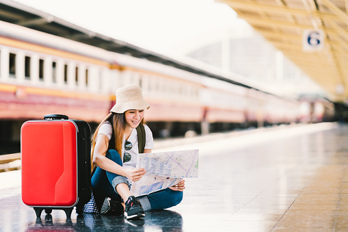 Asian backpack traveler woman using generic local map, siting alone at train station platform with luggage. Summer holiday travelling or young tourist concept