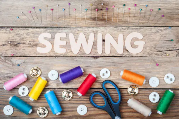 Photo of Sewing background with copy space. Colorful threads bobbins, word Sewing, scissors, pins and buttons on old wooden table. Accessories for handmade. Top view