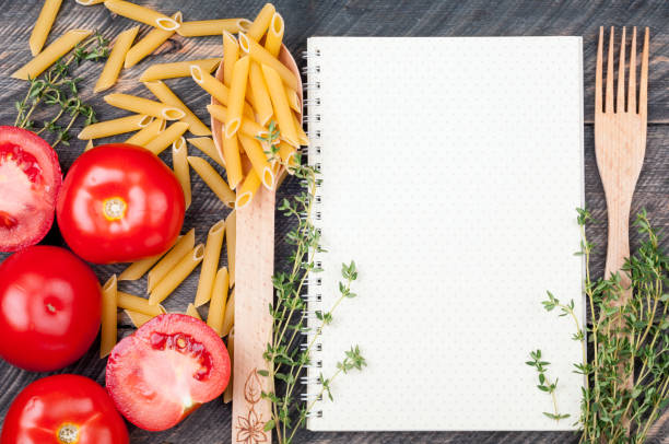 istock Open sheet cookbook with pattern polka dot, spoon with penne pasta, tomatoes, thyme on old wooden background. Ingredients for cooking. Open blank recipe book. Top view 692563500