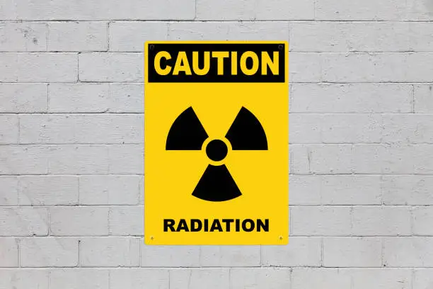 Yellow warning sign screwed to a brick wall to warn about a threat. In the middle of the panel, there is a biohazard symbol and the message is saying "Caution, Radiation".