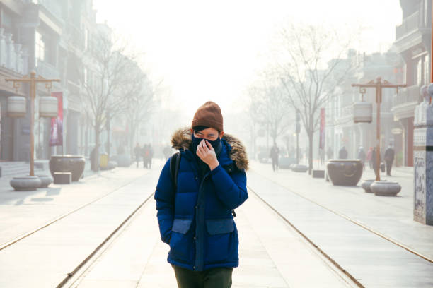 homem asiático andando e usando uma máscara de rosto em um dia nebuloso e nevoeiro poluição atmosférica como ele sofre de grave poluição em pequim, china - smog china beijing pollution - fotografias e filmes do acervo