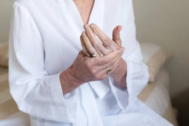 Close up cropped view of elderly woman in white bathrobe rubbing handcream into hands (selective focus)