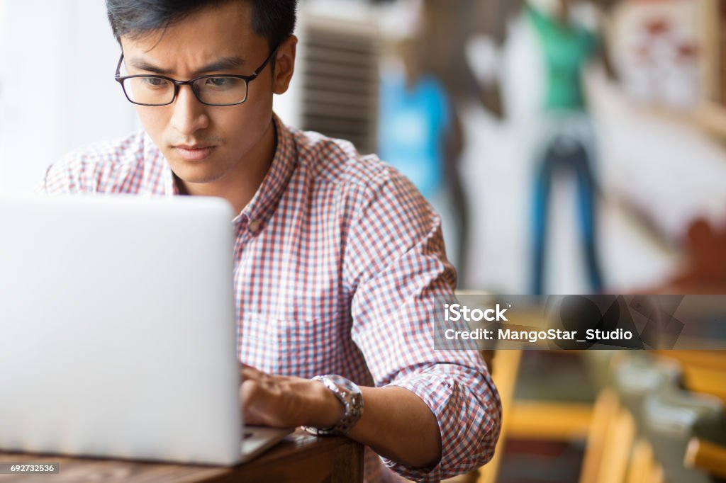 Serious young Asian student using laptop in cafe Portrait of serious young Asian student wearing glasses sitting at table and using laptop in cafe Serious Stock Photo