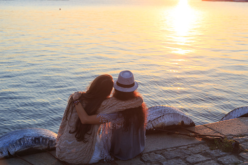 Rear view of two girlfriends enjoying summer vacations, sitting on beach and looking the sunrise. Embracing.