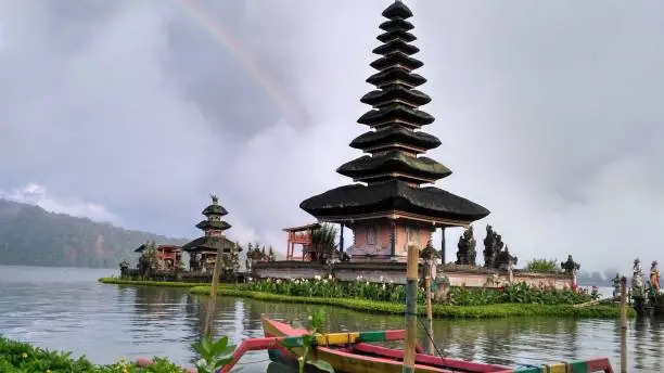 The view of the temple in the middle of the lake ulun danu in bali look very exotic, decorated with rainbow sky, traditional boat edge of the lake, and a calm atmosphere.