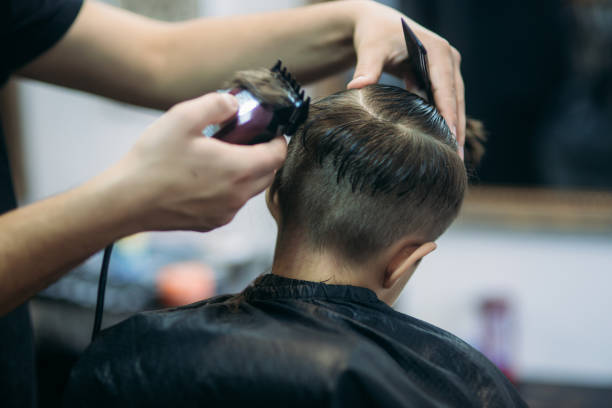 Little Boy Getting Haircut By Barber While Sitting In Chair At Barbershop. Little boy on a haircut in the barber sits on a chair. teenager boy haircuts stock pictures, royalty-free photos & images
