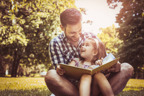 単一の父は小さな娘を草の上に座って、本の話を読んでします。少女は父の膝の上に座っています。 - family with two children family park child ストックフォトと画像