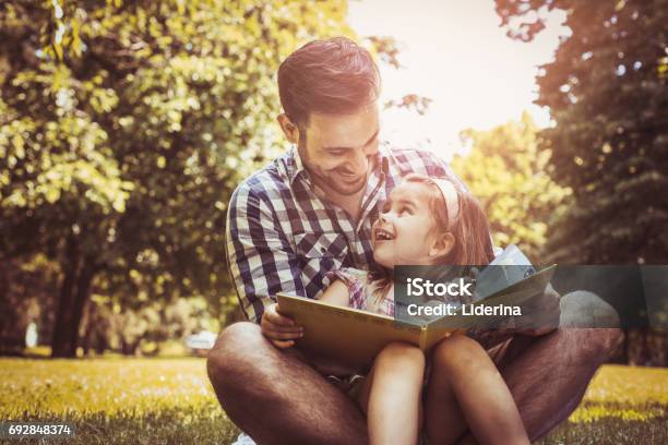 Single Father Sitting On Grass With Little Daughter And Reading Book Story Little Girl Sitting On Father Lap Stock Photo - Download Image Now