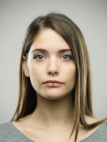 Close-up portrait of confident beautiful young woman with serene expression. Real people female is against gray background looking at camera. She has long brown hair. Vertical studio photography from a DSLR camera. Sharp focus on eyes.