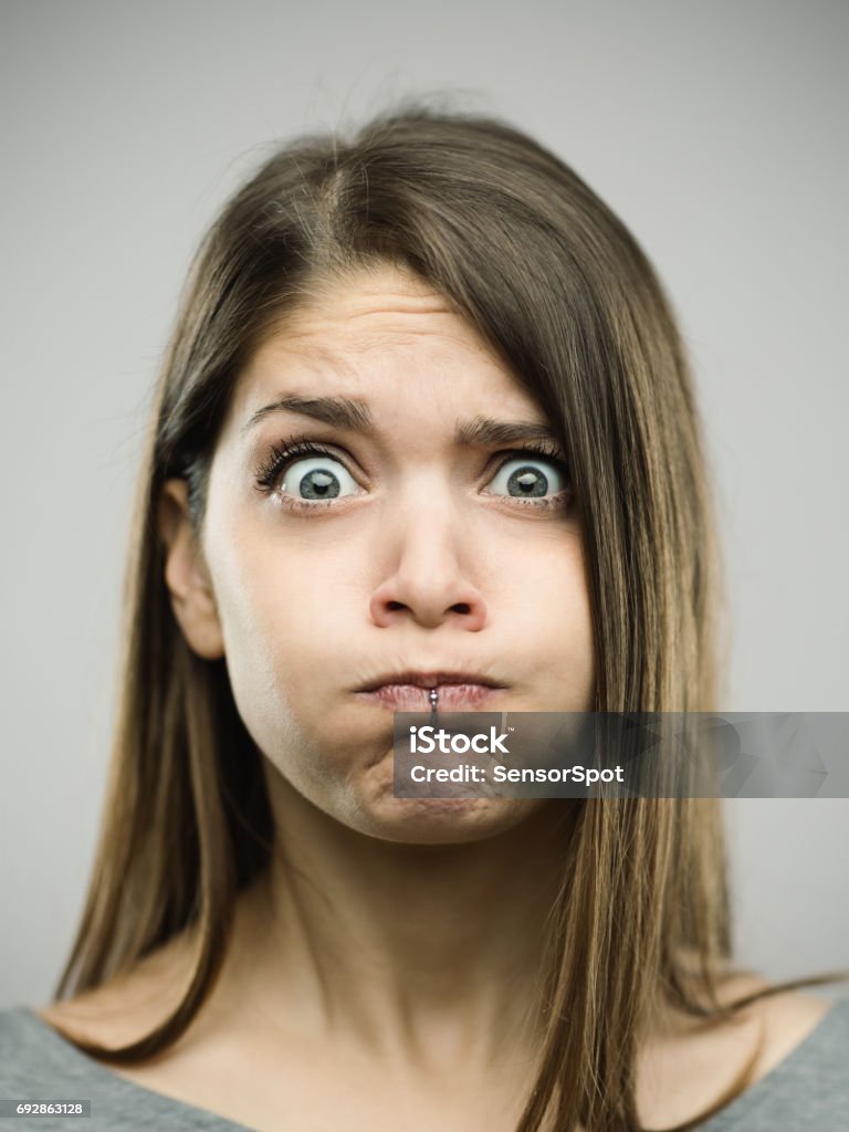 Real young woman with disgust expression Close-up portrait of disgusted beautiful young woman puffing cheeks. Real people female is against gray background with nausea expression. She has long brown hair. Vertical studio photography from a DSLR camera. Sharp focus on eyes. Holding Breath Stock Photo