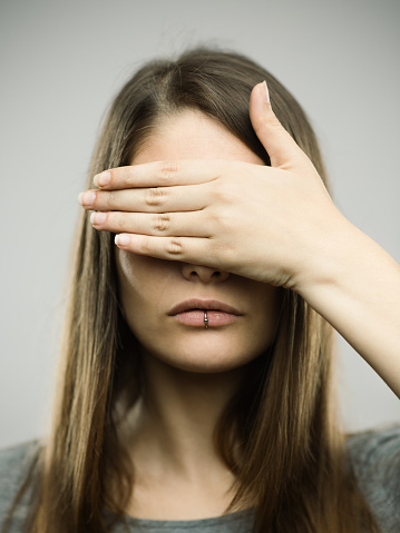 Close-up portrait of beautiful young woman covering her eyes with the hand. Real people female is against gray background with see no evil gesture. She has long brown hair. Vertical studio photography from a DSLR camera. Sharp focus on eyes.