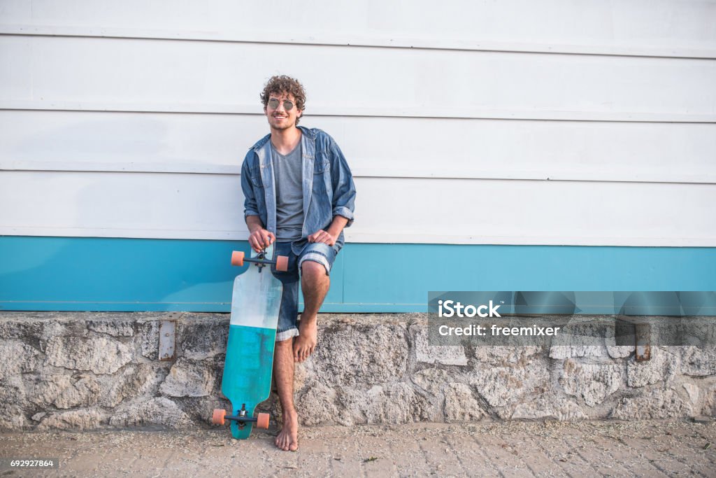 Funky skaterboy Young beautiful hipster man leaning on the wall near the beach with longboard next to him Men Stock Photo