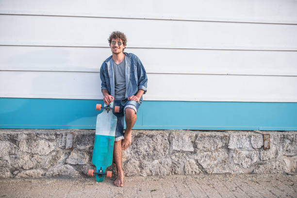 Funky skaterboy Young beautiful hipster man leaning on the wall near the beach with longboard next to him longboarding stock pictures, royalty-free photos & images