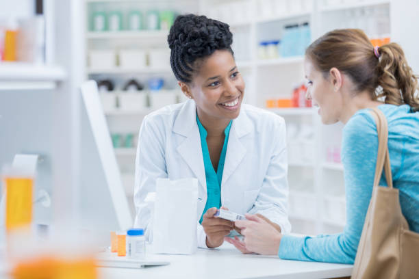 Pharmacy customer asks pharmacist question about medication Mid adult female Caucasian pharmacy customer asks African American female pharmacist question about over the counter cold and flu medication. chemist stock pictures, royalty-free photos & images