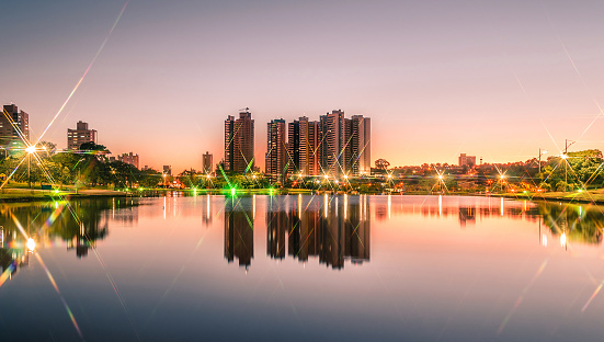 Sunset at a lake of a park with the city on background. Scene reflected on water. Landscape of a beautiful warm sunset at Parque das Nacoes Indigenas in Campo Grande, Brazil.