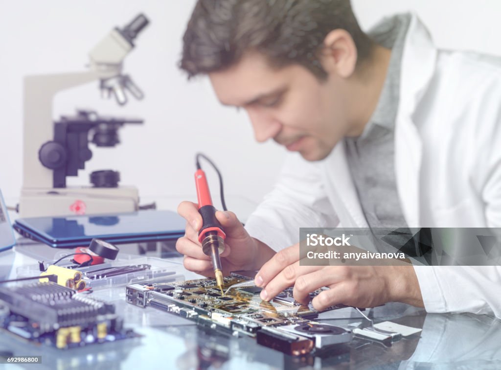 Young energetic male tech or engineer repairs electronic equipment Electronics background. Young energetic male tech or engineer repairs electronic equipment in research facility. Shallow DOF, focus on the hands of the worker. This image is toned. Circuit Board Stock Photo