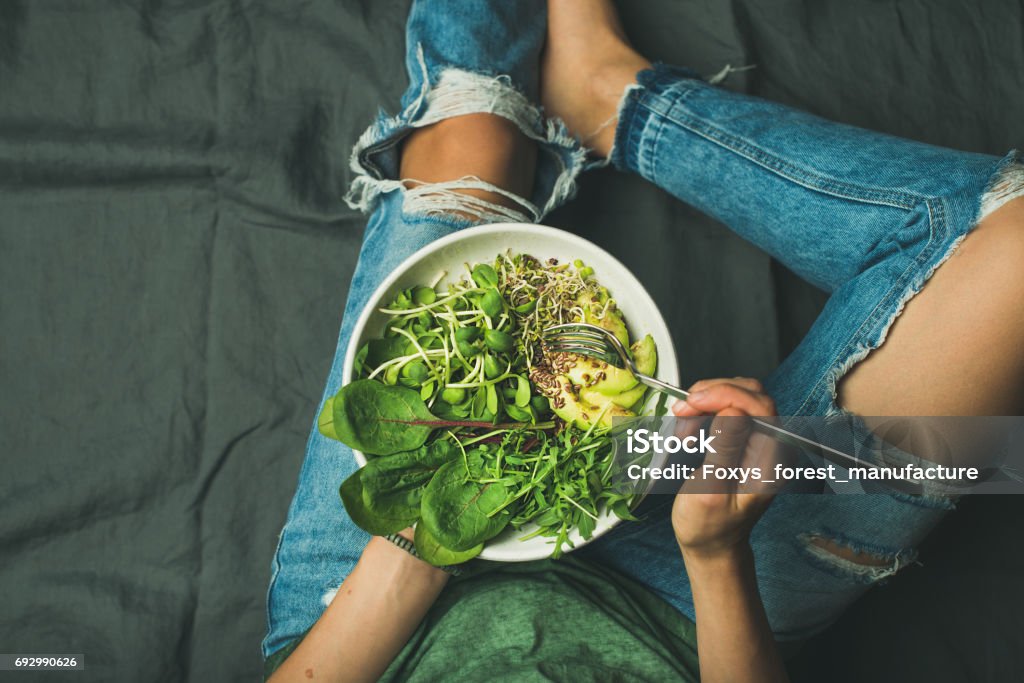 Vegetarian breakfast bowl with spinach, arugula, avocado, seeds and sprouts Green vegan breakfast meal in bowl with spinach, arugula, avocado, seeds and sprouts. Girl in jeans holding fork with knees and hands visible, top view, copy space. Clean eating, vegan food concept Eating Stock Photo