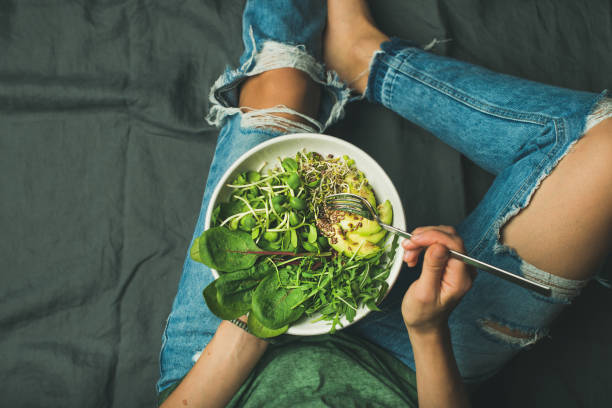 Vegetarian breakfast bowl with spinach, arugula, avocado, seeds and sprouts Green vegan breakfast meal in bowl with spinach, arugula, avocado, seeds and sprouts. Girl in jeans holding fork with knees and hands visible, top view, copy space. Clean eating, vegan food concept foxys_forest_manufacture stock pictures, royalty-free photos & images