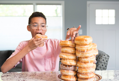 A boy eats a donut and looks at the huge pile of donuts in front of him.  He is hispanic and has glasses and a pink shirt.  He is sitting at a kitchen counter inside a home.