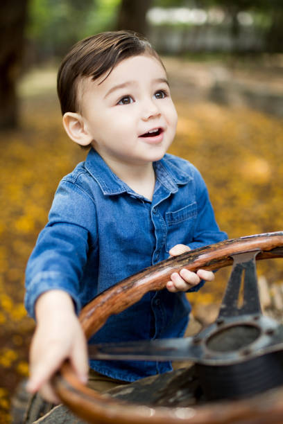 A little latin boy holding a steering wheel, looking up and smiling in a vertical medium shot outdoors.