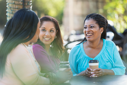 Group of three multi-ethnic women sitting around an outdoor table on a patio drinking coffee and conversing.  The woman in purple is mixed race African-American. The focus is on her African-American friend wearing blue.