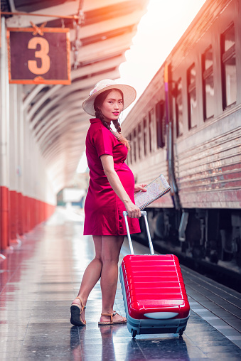 Asian pregnant woman in red dress carrying red luggage and map walking at railway station