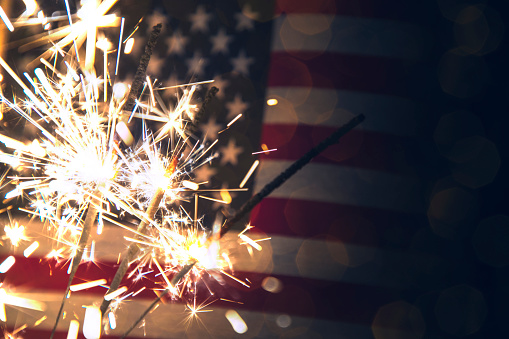 A stock photo of a USA stars and stripes flag with sparklers in the foreground. Perfect for designs and articles about the 4th of July and the USA.