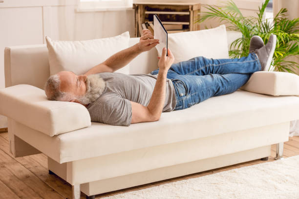 istock senior bearded man using digital tablet and lying on sofa at home 693205202