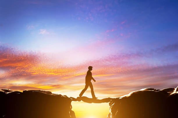 Man walking over precipice between mountains, another man being a bridge