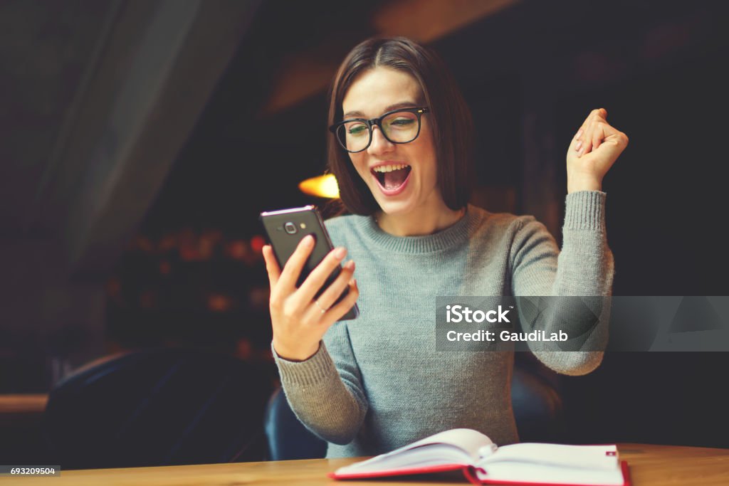 Emotional young attractive female expressing surprise Checking email box and reading messages with congratulation of winning using modern smartphone connected to 4G internet in coworking office Giving Stock Photo