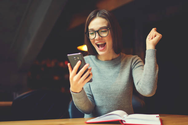 Emotional young attractive female expressing surprise Checking email box and reading messages with congratulation of winning using modern smartphone connected to 4G internet in coworking office Best Student Apps stock pictures, royalty-free photos & images