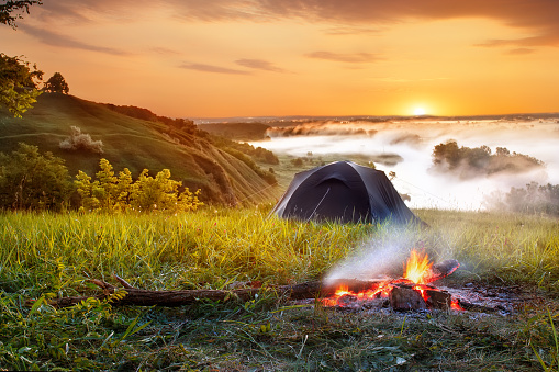 campfire and tent at sunrise. View of tourist tent on green meadow. Camping background. Adventure travel active lifestyle freedom concept. Summer landscape. Foggy meadow in the sunny beams
