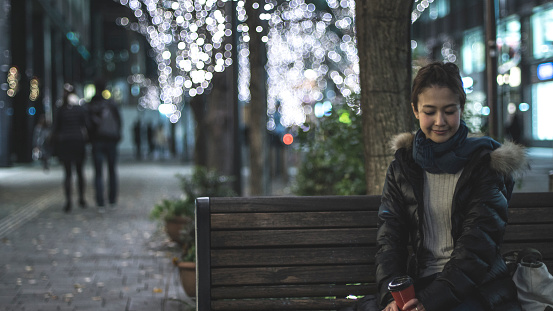 A young woman is sitting on a bench alone in a town at night. Street trees in the town are illuminated with many small white lights in winter season. She is looking down and resting there.
