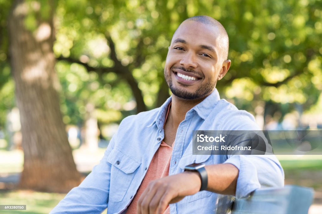 Happy african american man Young african man relaxing at park in a summer day. Happy black cheerful guy feeling good and sitting on bench at park. Smiling american man looking at camera outdoor. Men Stock Photo