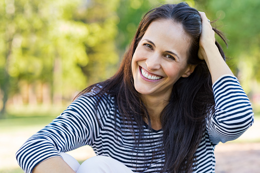 Happy latin woman having fun at park while touching hair. Hispanic woman enjoying holiday while sitting on grass in park. Portrait of smiling mature woman relaxing and looking at camera.
