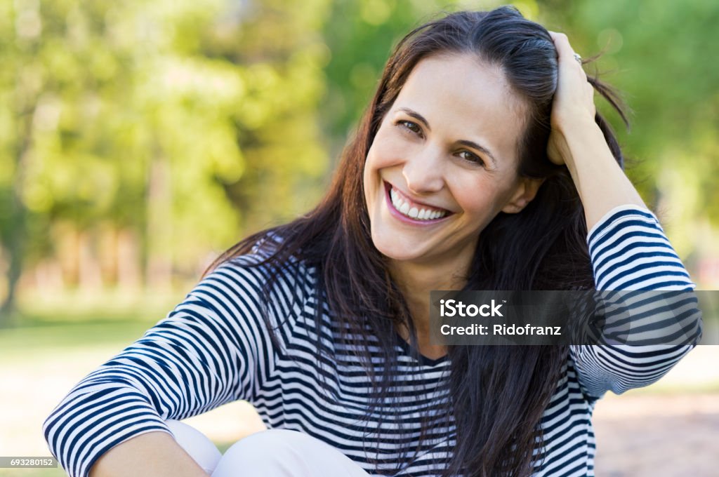 Laughing mid woman at park Happy latin woman having fun at park while touching hair. Hispanic woman enjoying holiday while sitting on grass in park. Portrait of smiling mature woman relaxing and looking at camera. Women Stock Photo